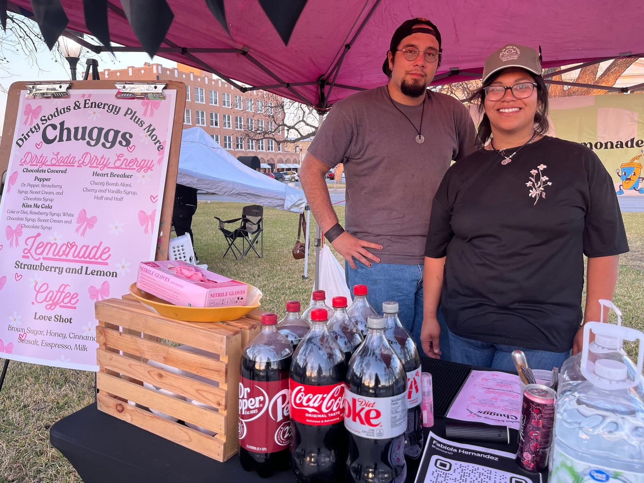 A picture of a couple standing underneath a pink tent, a table in front of them with eight soda bottles, a wooden crate, two jugs of water, and a printed menu for "Chuggs". To the left of them is a larger printed version of their menu, advertising their Dirty Soda, Dirty Energy, Lemonade, and Coffee