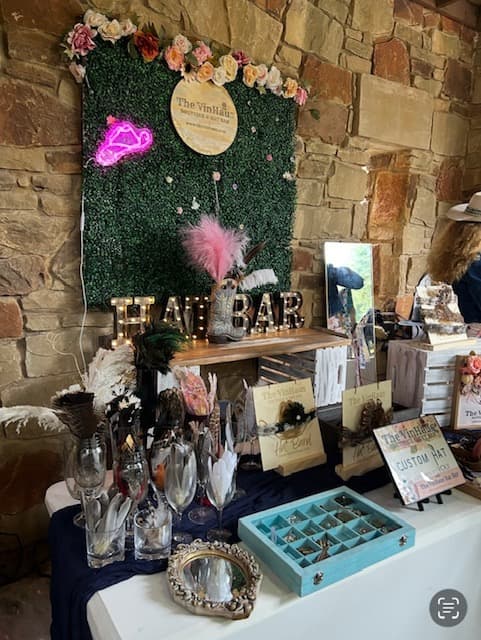 an image on the Vinhuas Hat Bar set up in front of a stone wall. Feathers and hat bands are displayed on a table. Behind, on another table, are the words "Hat Bar" in light-up letters, as well as a cowboy boot with a pink feather coming out of it