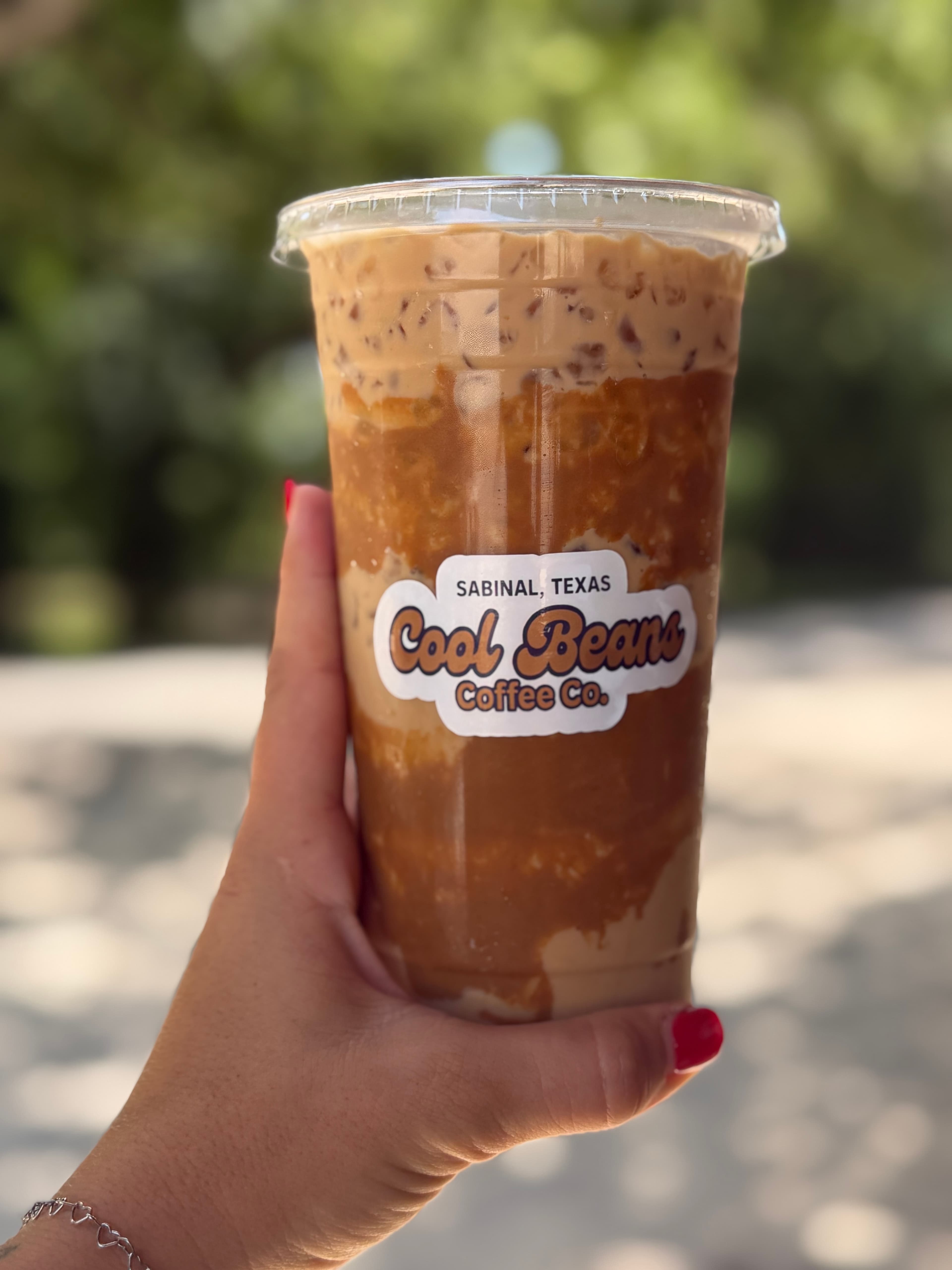 A hand holding an iced coffee in a clear cup with "Cool Beans Coffee Co., Sabinal, Texas" on the sticker of the cup
