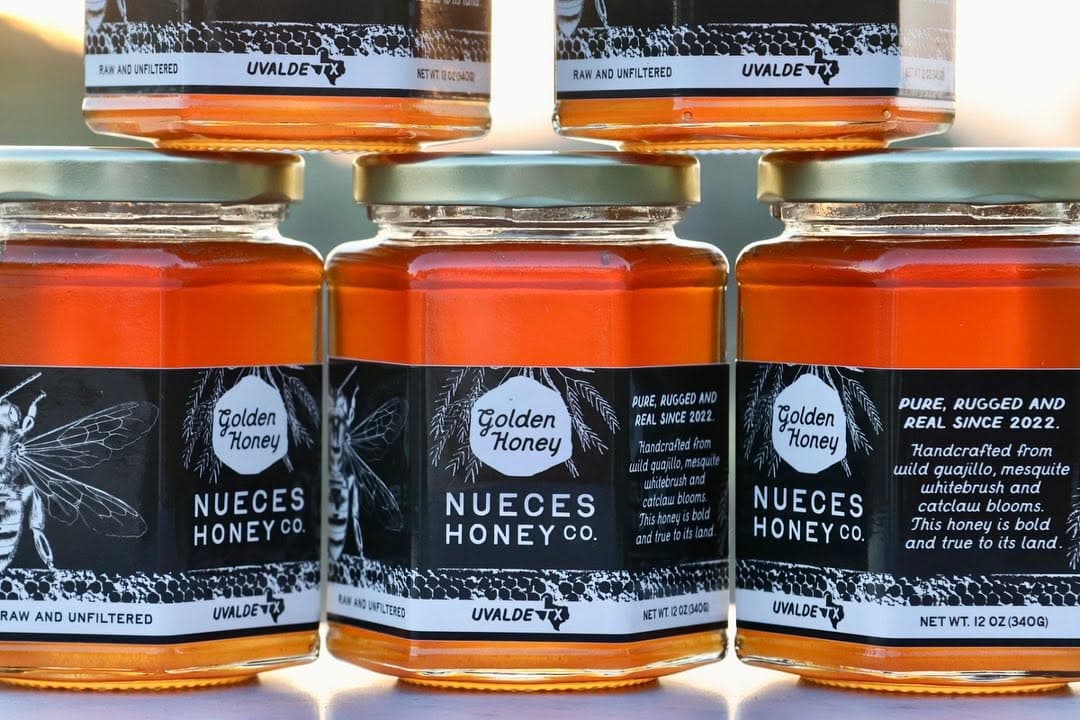 Five jars of golden local honey stacked and displayed. the label reading "Golden Honey. Nueces Honey Co. Raw and unfliteres. Uvalde, Texas. Pure, rugged and real since 2022. Handcrafted from wild guajillo, mesquite, whitebrush and catclaw blooms. This honey is bold and true to its land. NET WT 12 OZ (340G)